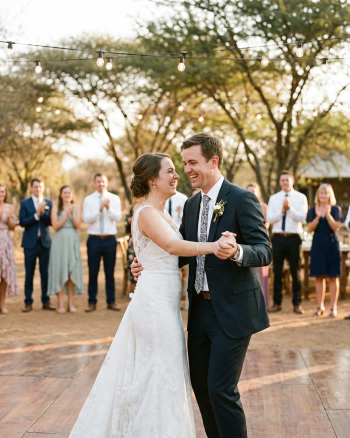 A bride and groom laughing mid-dance at golden hour while guests clap behind them